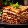 Ein Stück klassische Lasagne mit mehreren Schichten aus Nudelplatten, würziger Bolognese-Sauce, cremiger Béchamel und geschmolzenem Käse, serviert auf einem schwarzen Teller, garniert mit frischem Basilikum